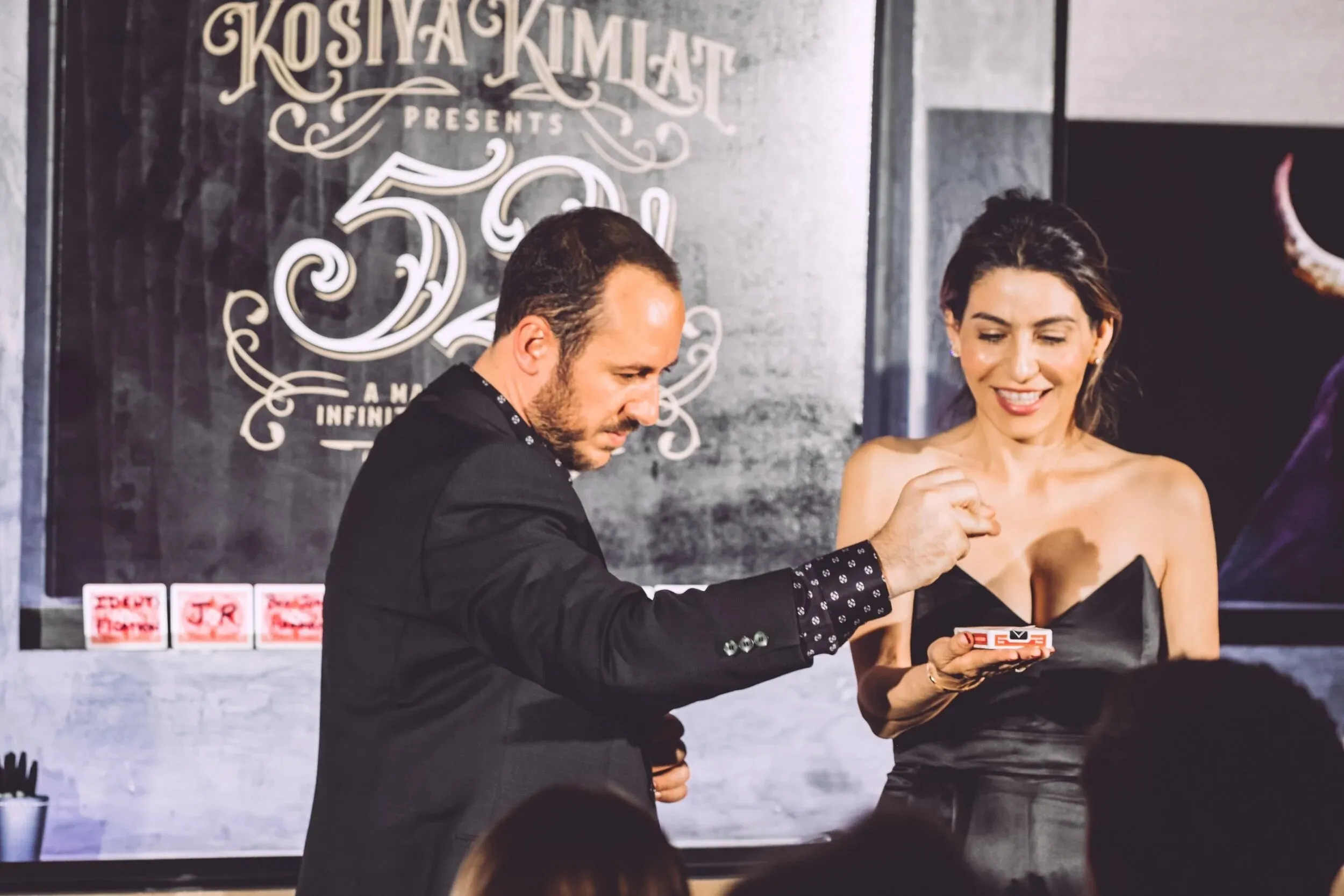 Kostya Kimlat performing a card trick for a guest at 52!, the show's artwork on the wall behind them