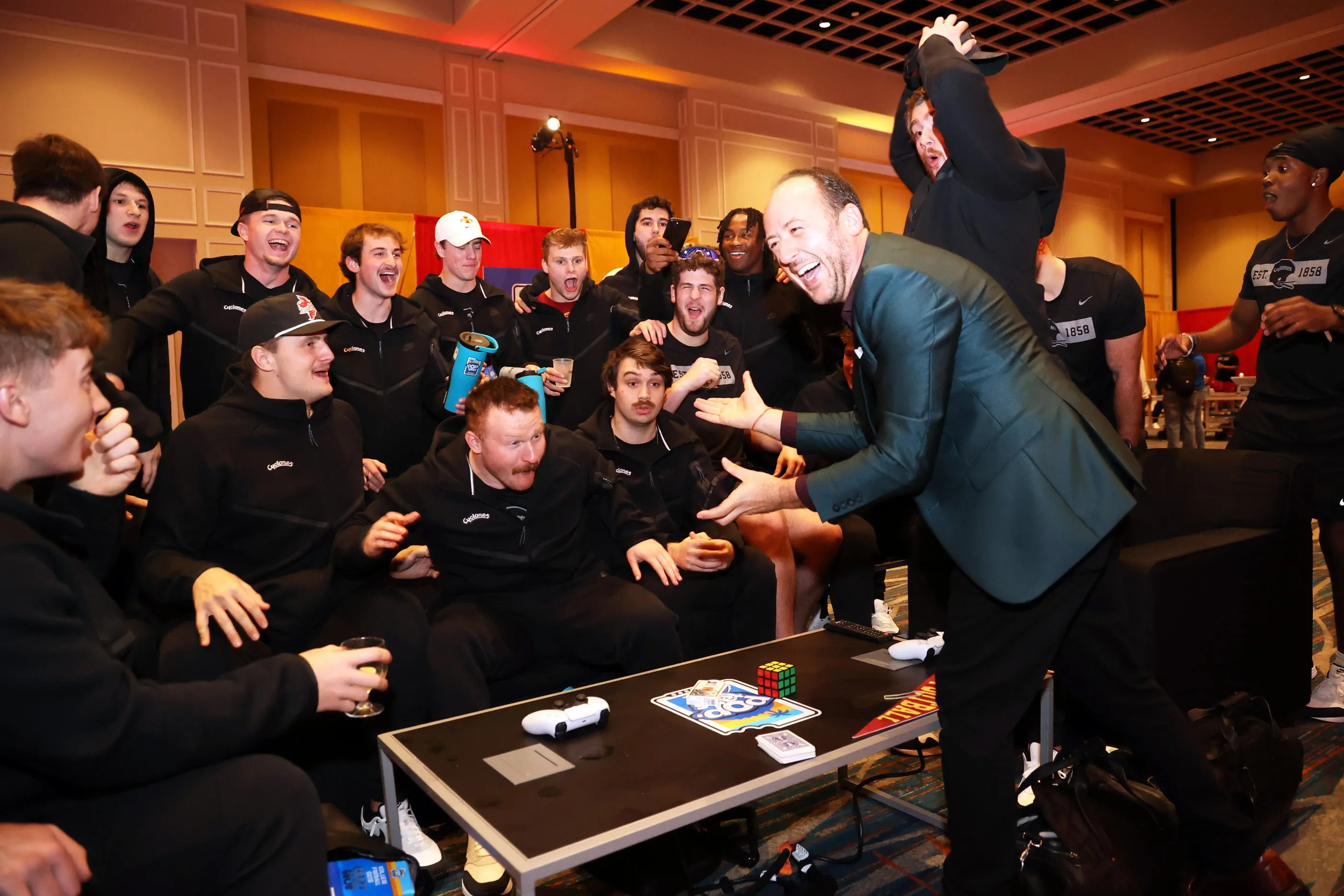 A college football team crowded around a low table, all of them laughing and reaching forward, as Kostya Kimlat performs close-up magic