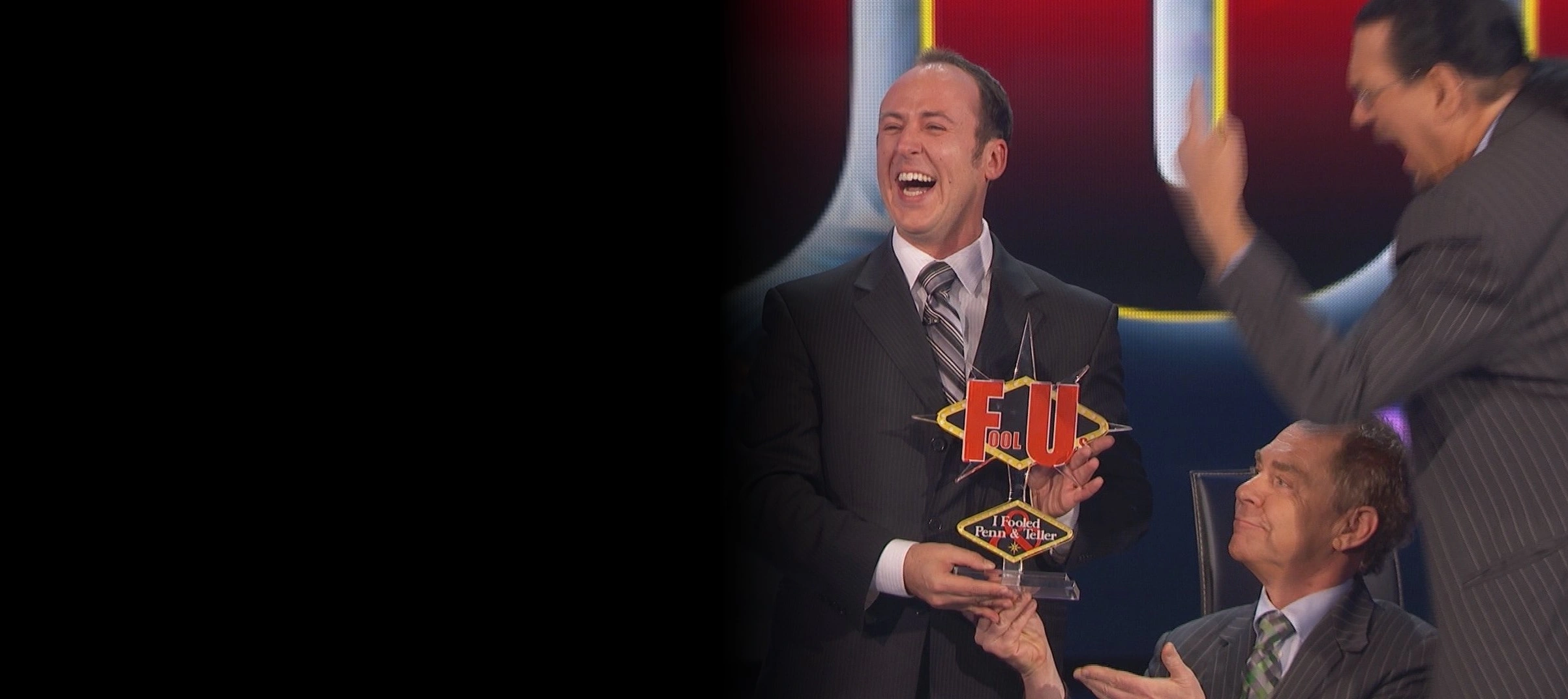 Kostya Kimlat holding the Fool Us trophy on stage as Penn Jillette and Teller applaud