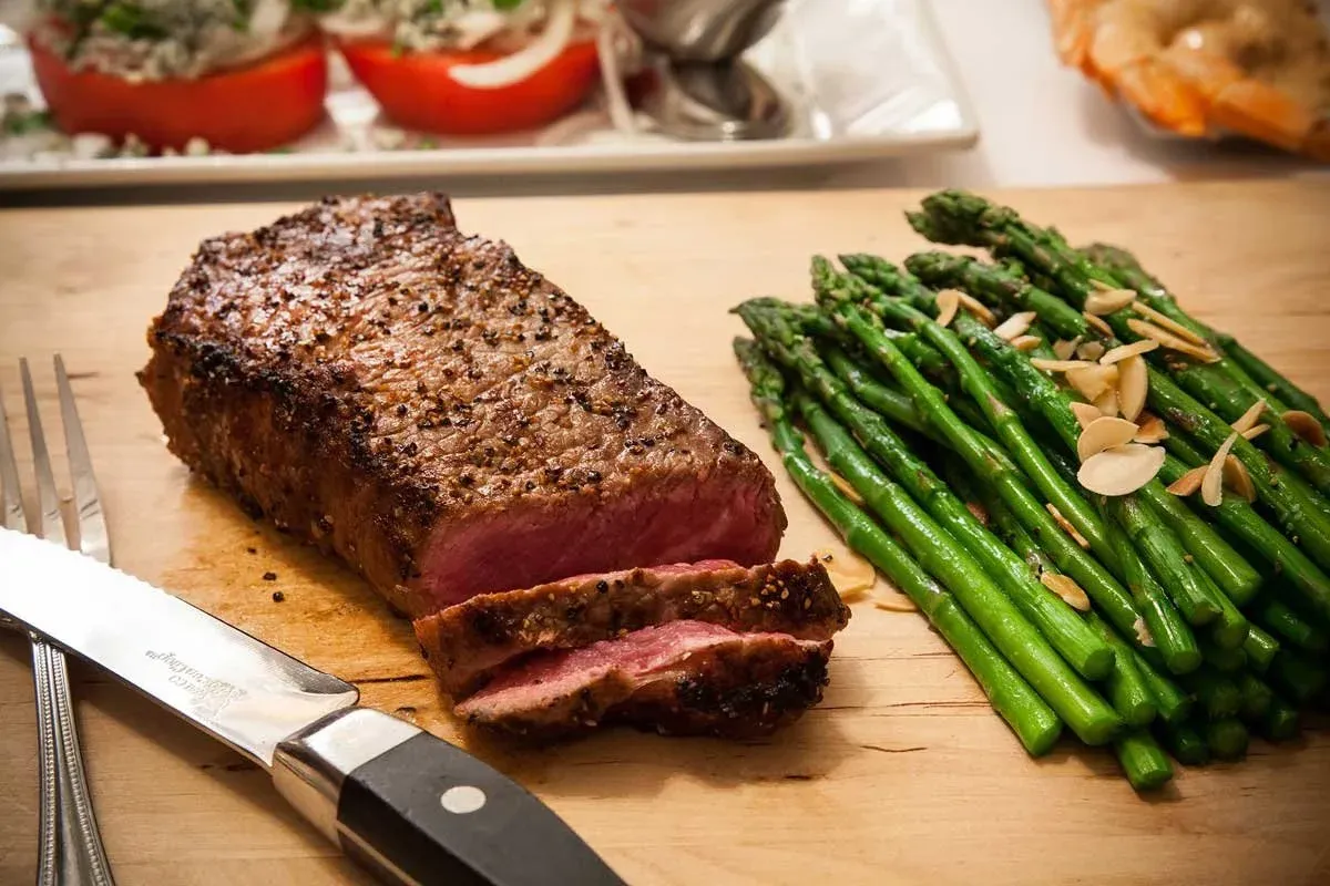 A sliced prime strip steak on a carving board beside a serving of asparagus with almonds