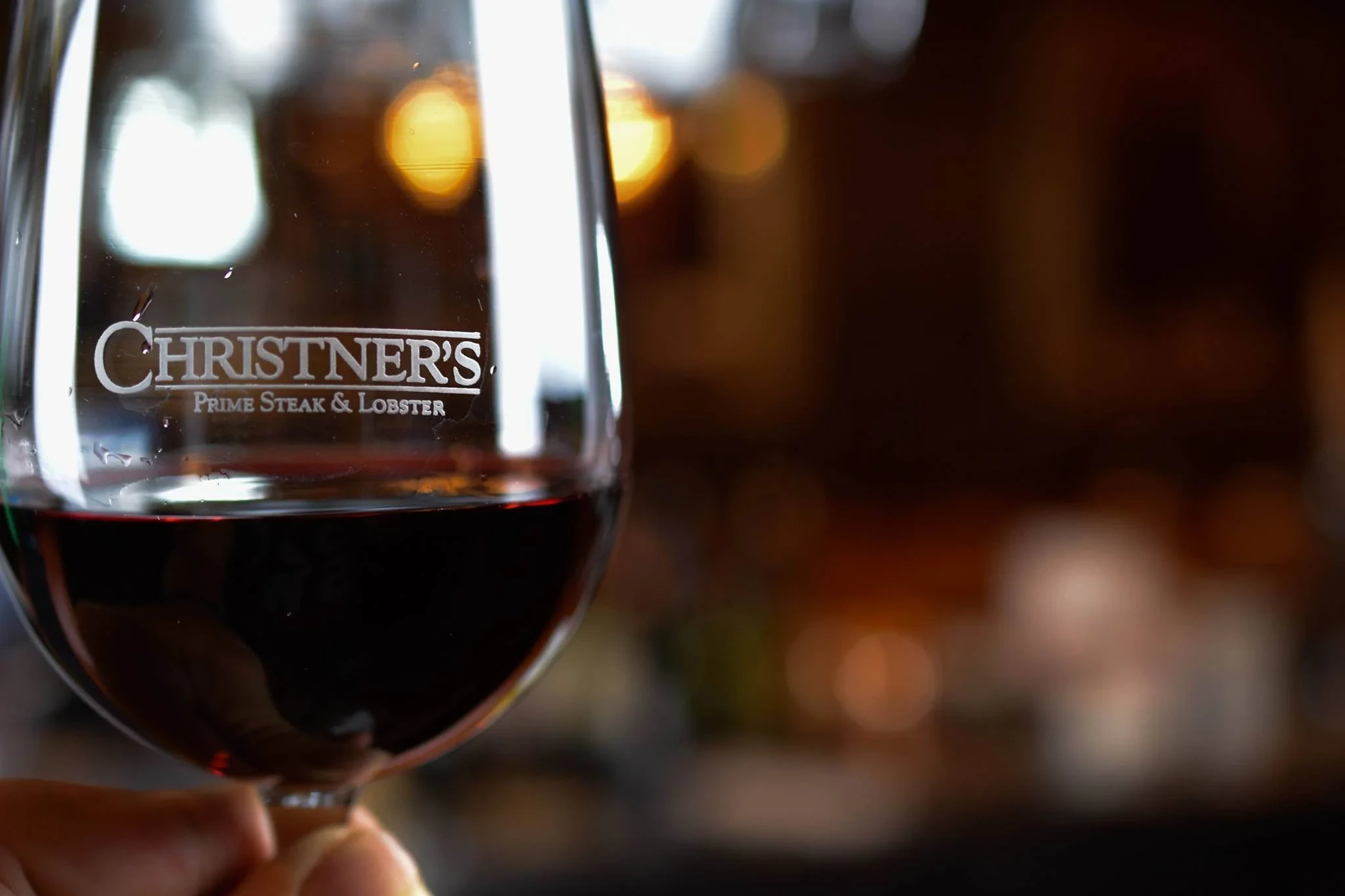 A glass of red wine engraved with the Christner’s Prime Steak & Lobster crest, held up in the warm light of the dining room