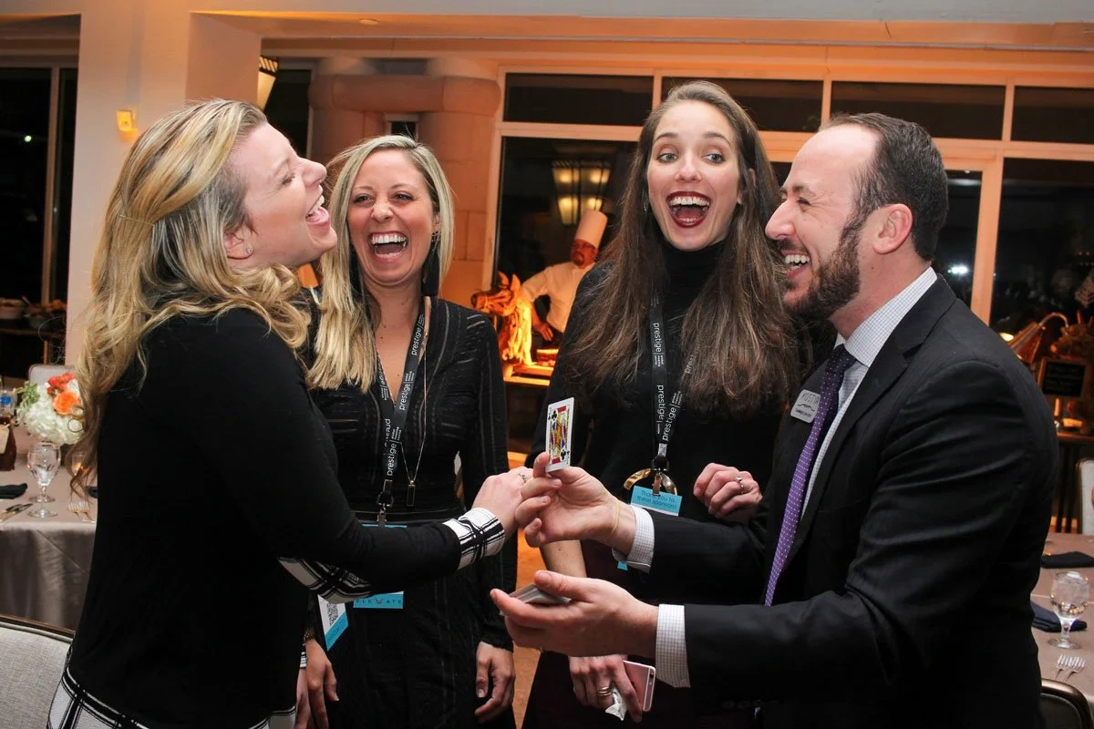 Three guests at a reception laughing as Kostya Kimlat holds up a chosen playing card between them