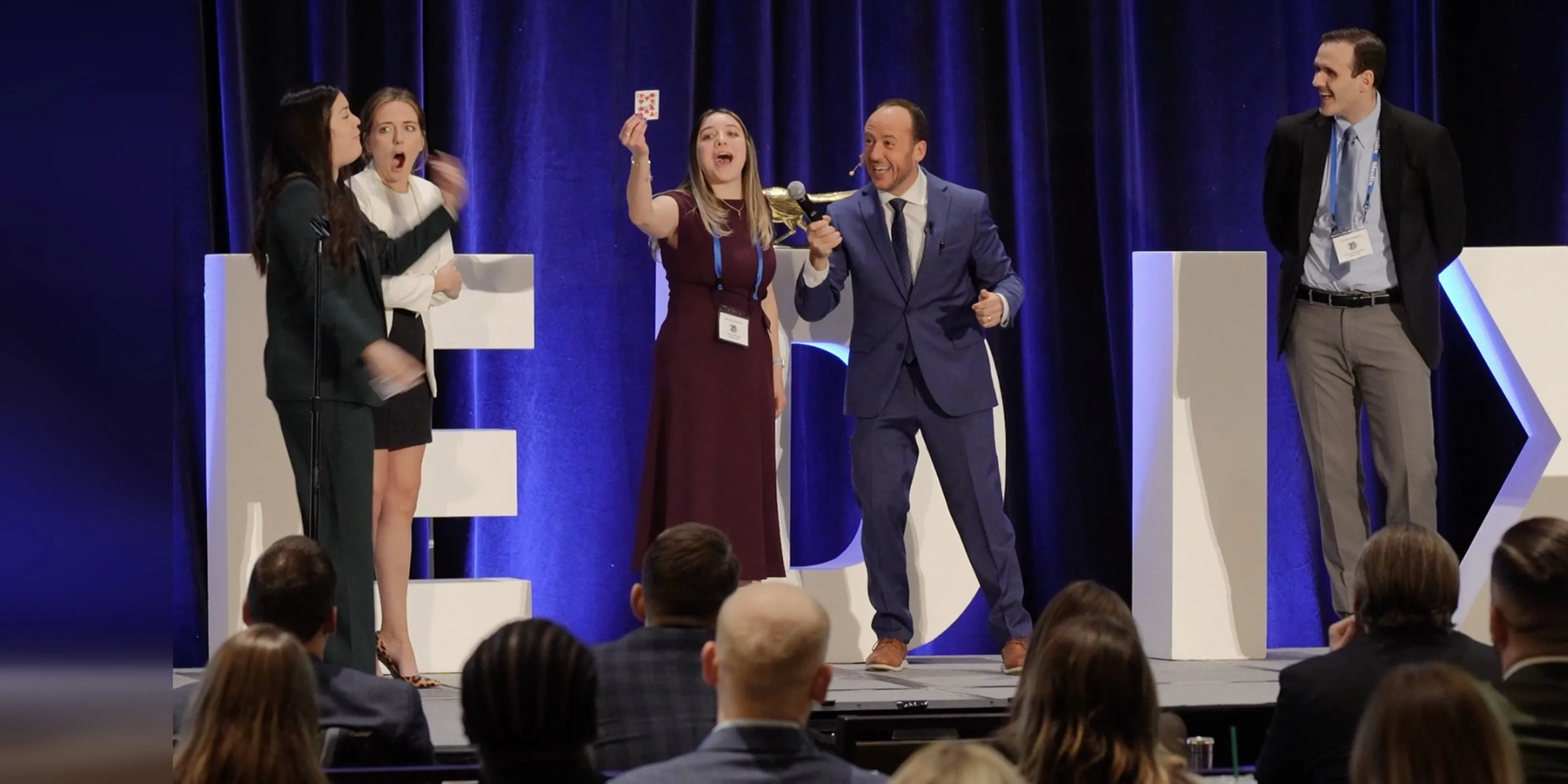 Conference volunteers on stage reacting, one holding up her chosen card, as Kostya Kimlat speaks into a microphone beside her