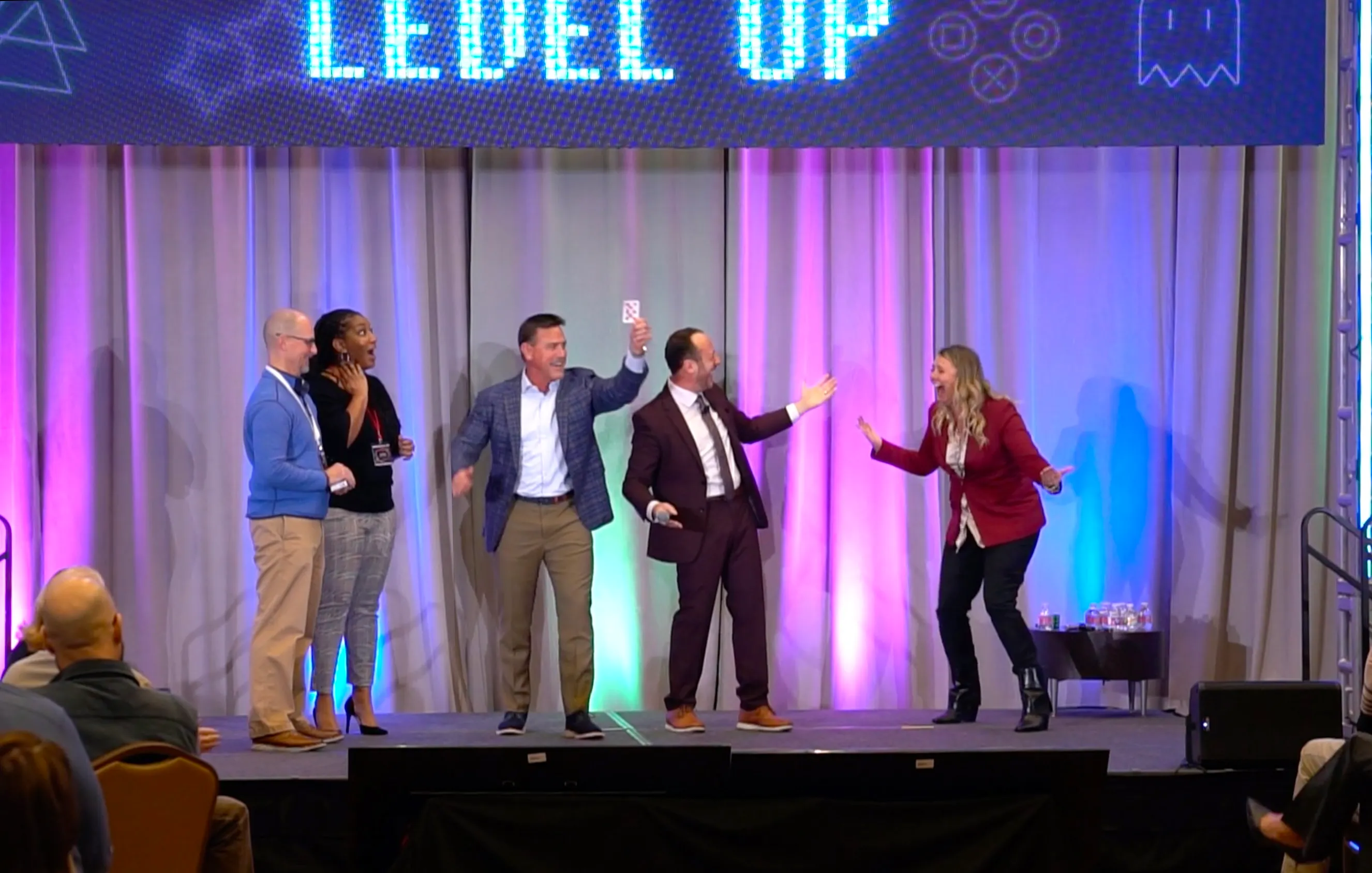 Volunteers on a conference stage laughing and throwing their hands up as one holds a playing card overhead, Kostya Kimlat beside them with a microphone