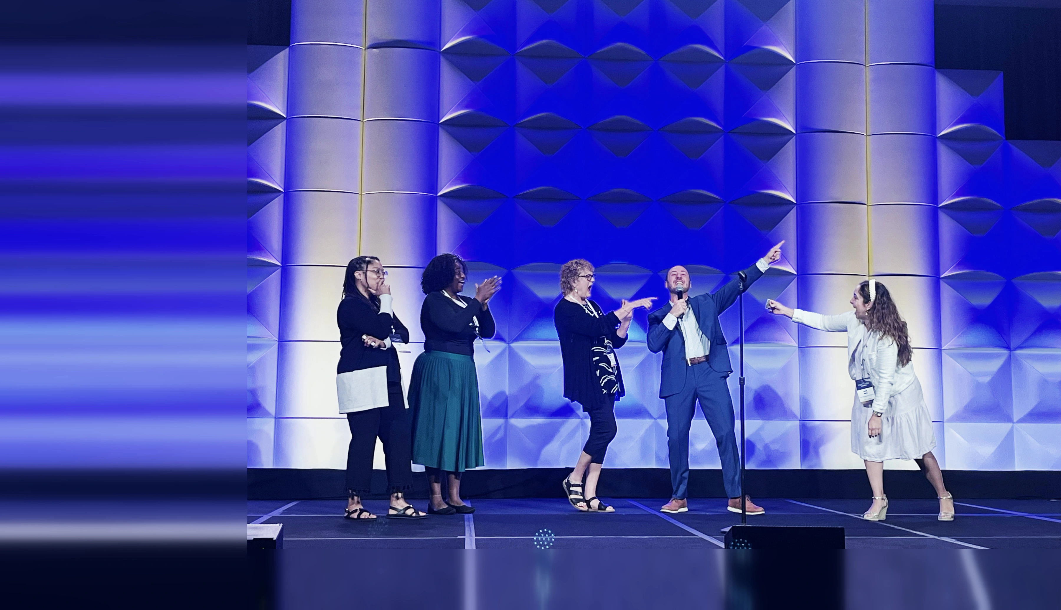 Four volunteers react on stage, one covering her mouth, one applauding, as Kostya Kimlat points past them, microphone in hand