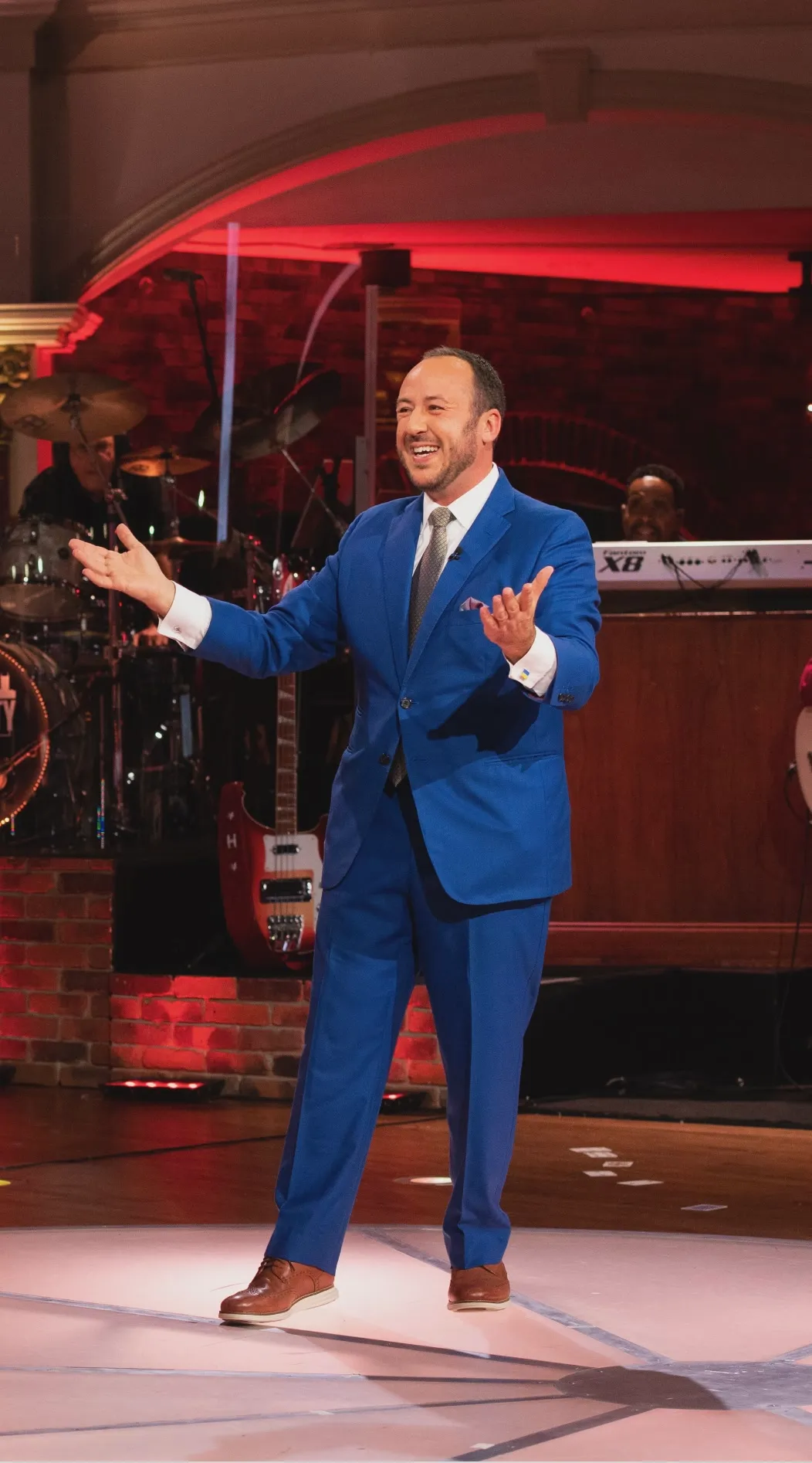 Kostya Kimlat mid-performance in a blue suit, arms open to the room, a live band on the stage behind him