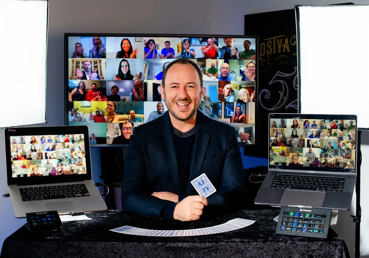 Kostya Kimlat in a broadcast studio holding a playing card, three screens around him filled with grids of guests watching