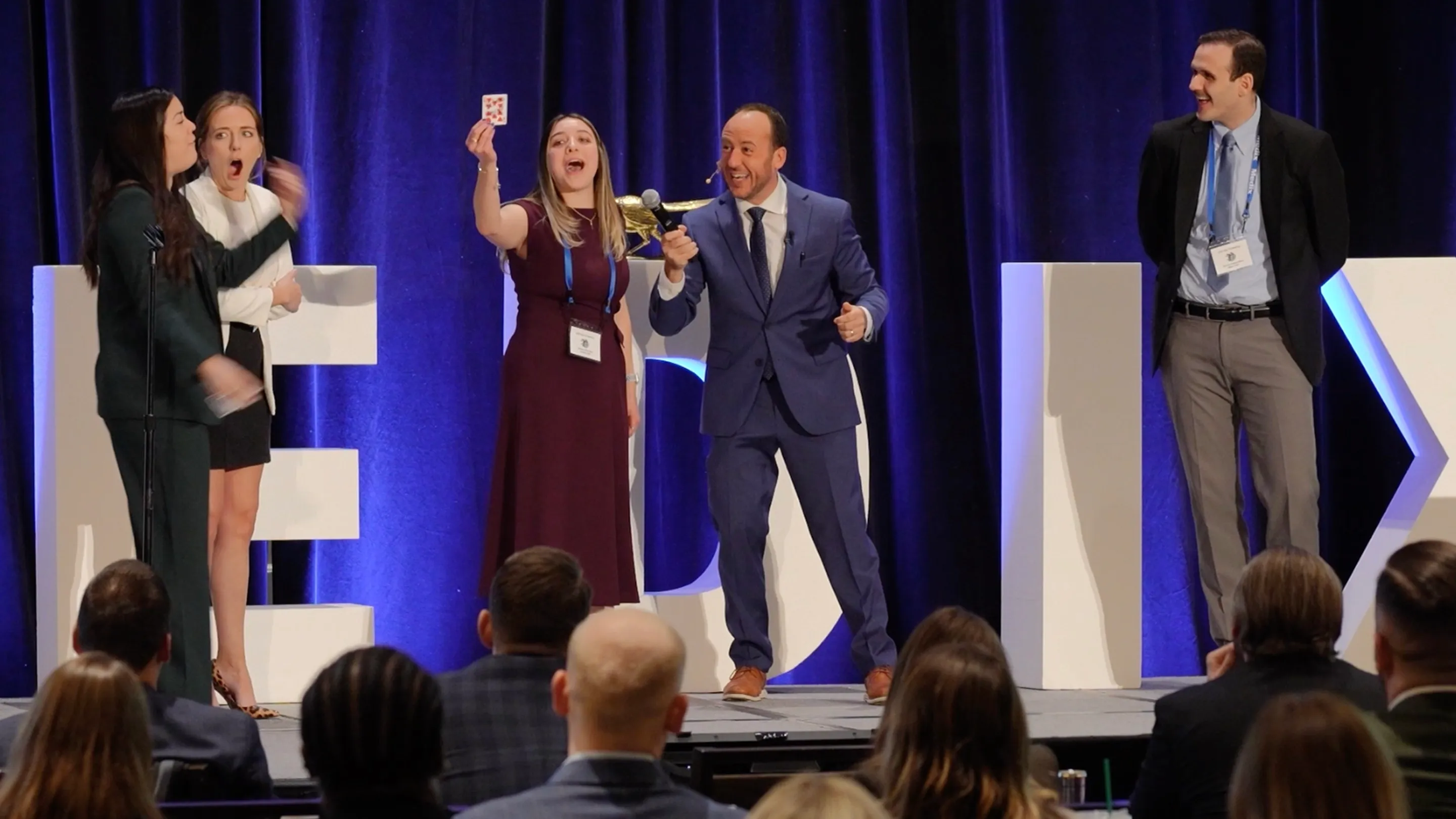 A conference attendee holds up her signed card, amazed, as Kostya reacts beside her on stage