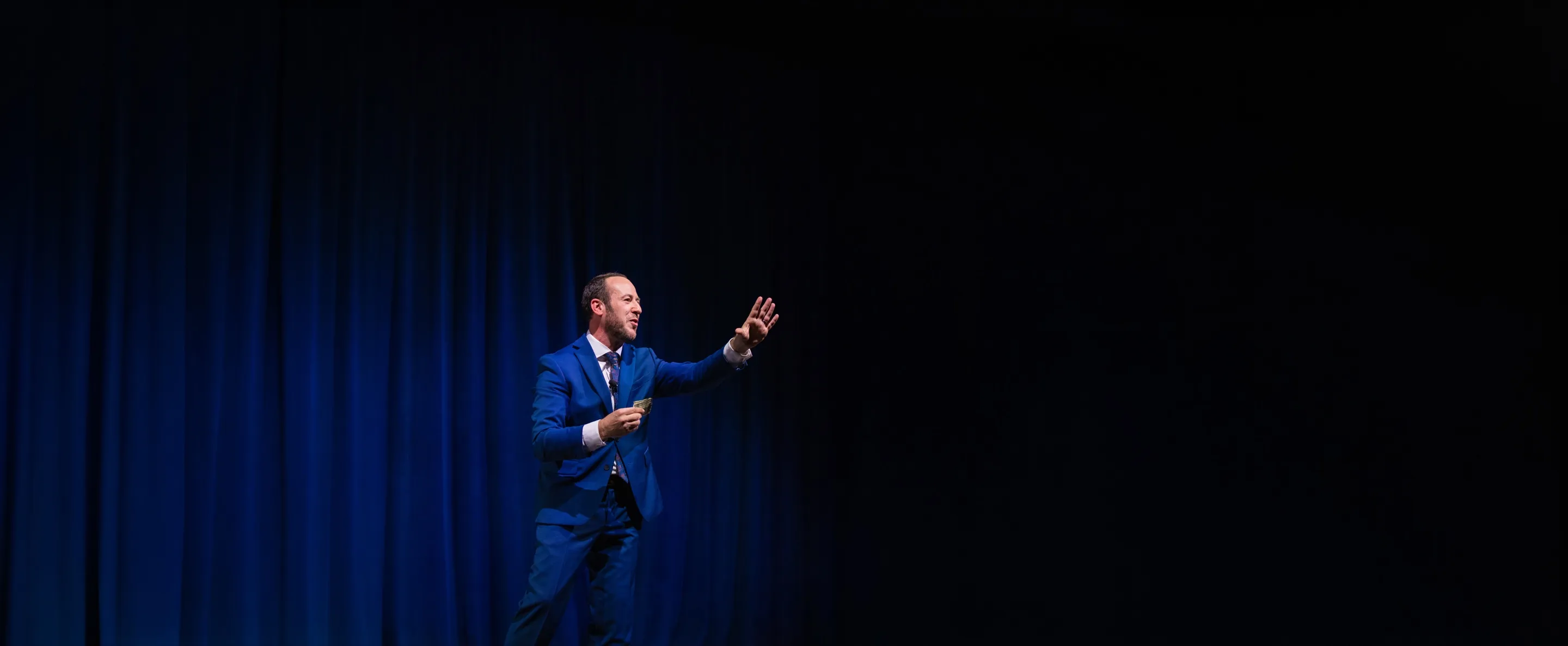Kostya Kimlat alone in a spotlight on a dark stage, one hand raised toward the audience