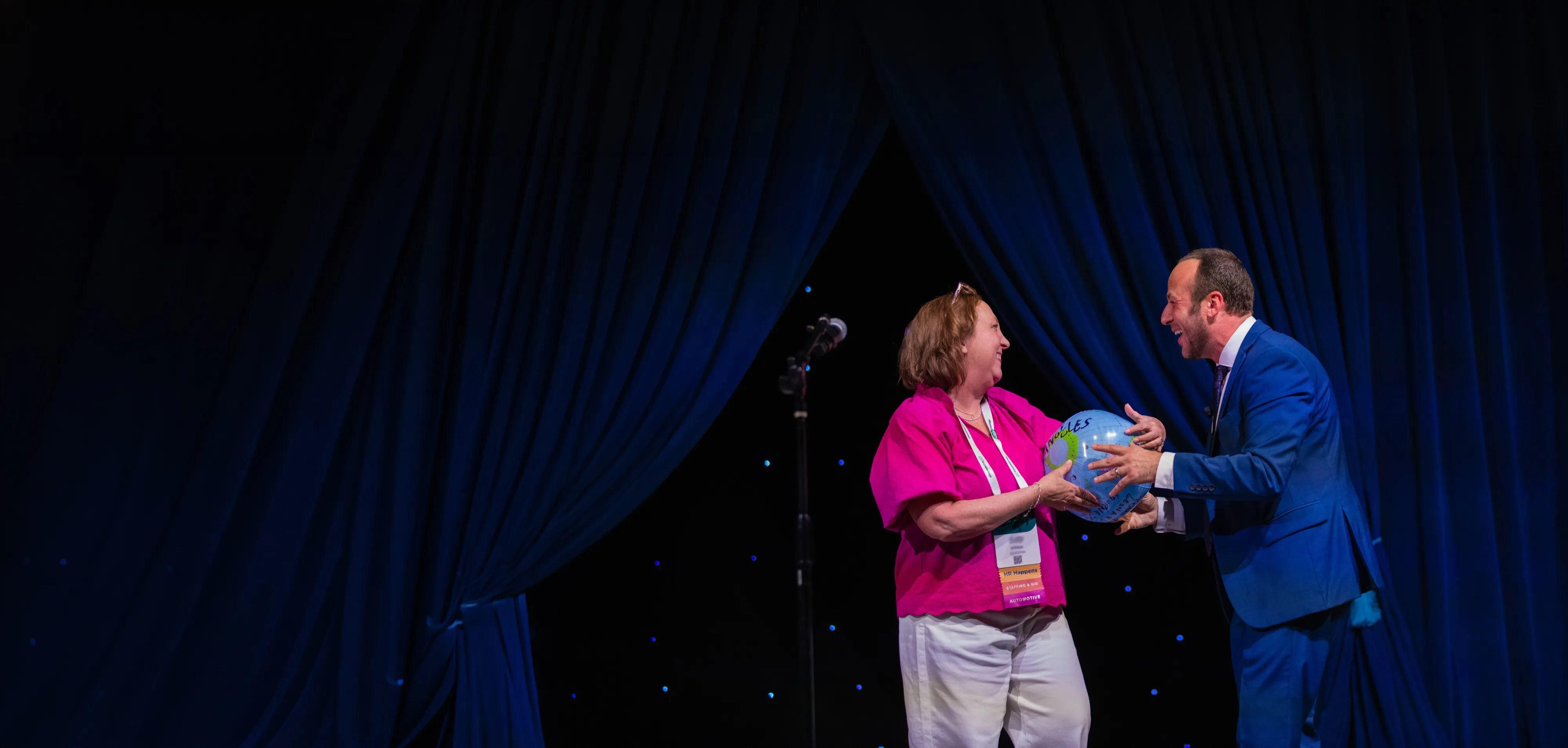 Kostya and an audience volunteer laughing together on stage as they hold an inflatable globe