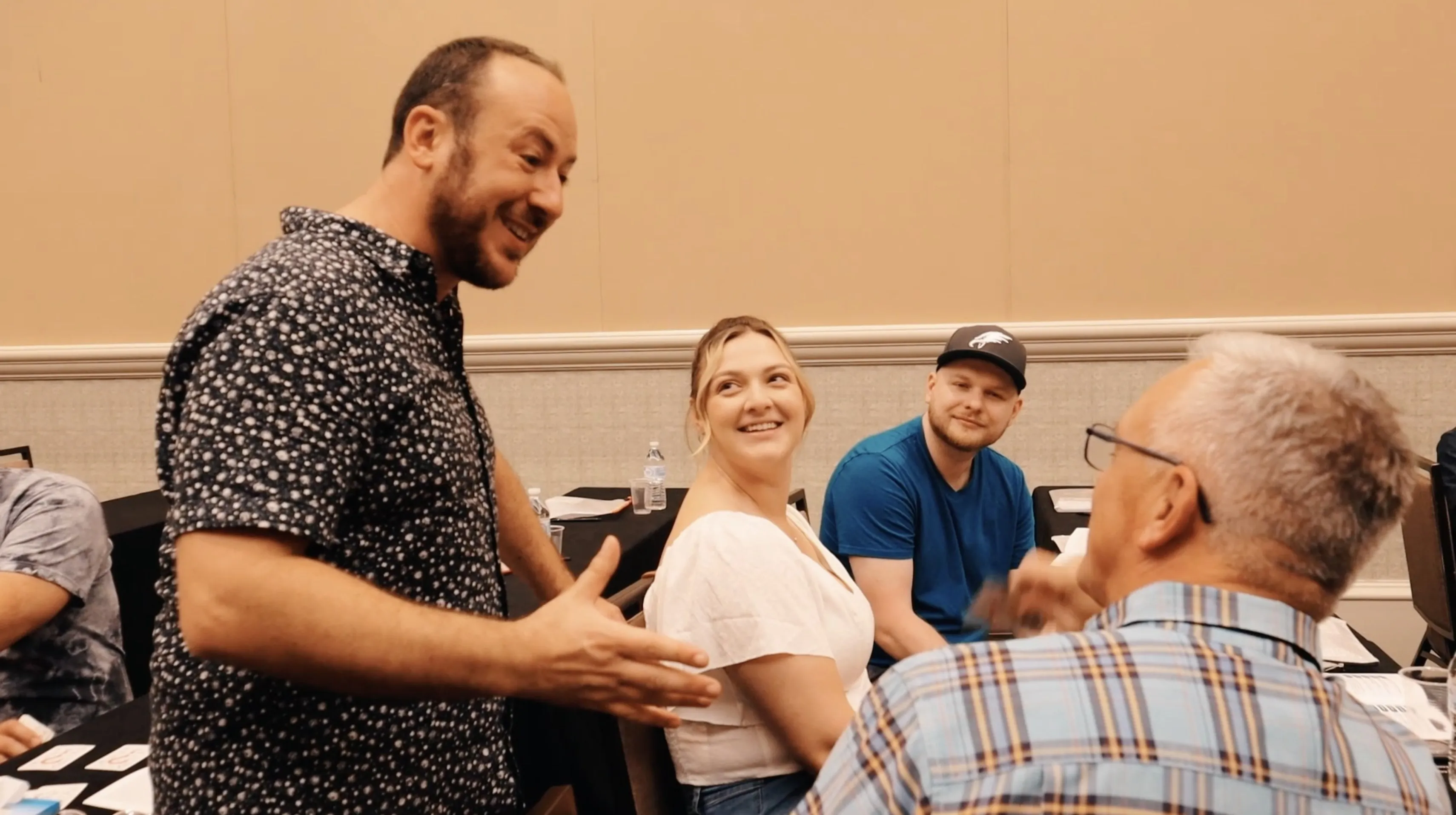 Kostya Kimlat standing at a table in a mentalism workshop, explaining something to attendees who smile up at him