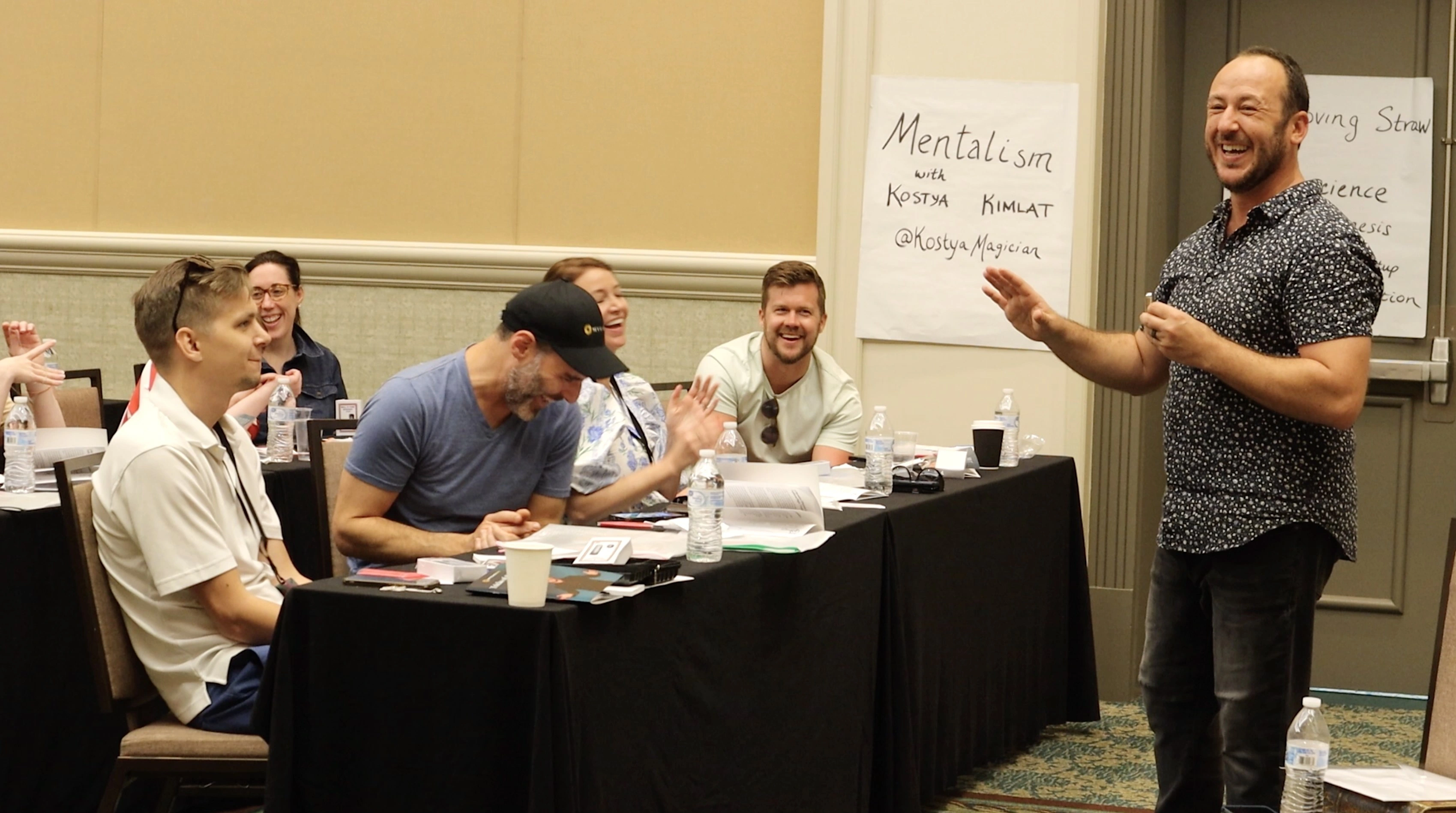 Kostya Kimlat laughing at the front of a workshop room as attendees at classroom tables laugh and applaud, a flip chart behind him reading Mentalism with Kostya Kimlat
