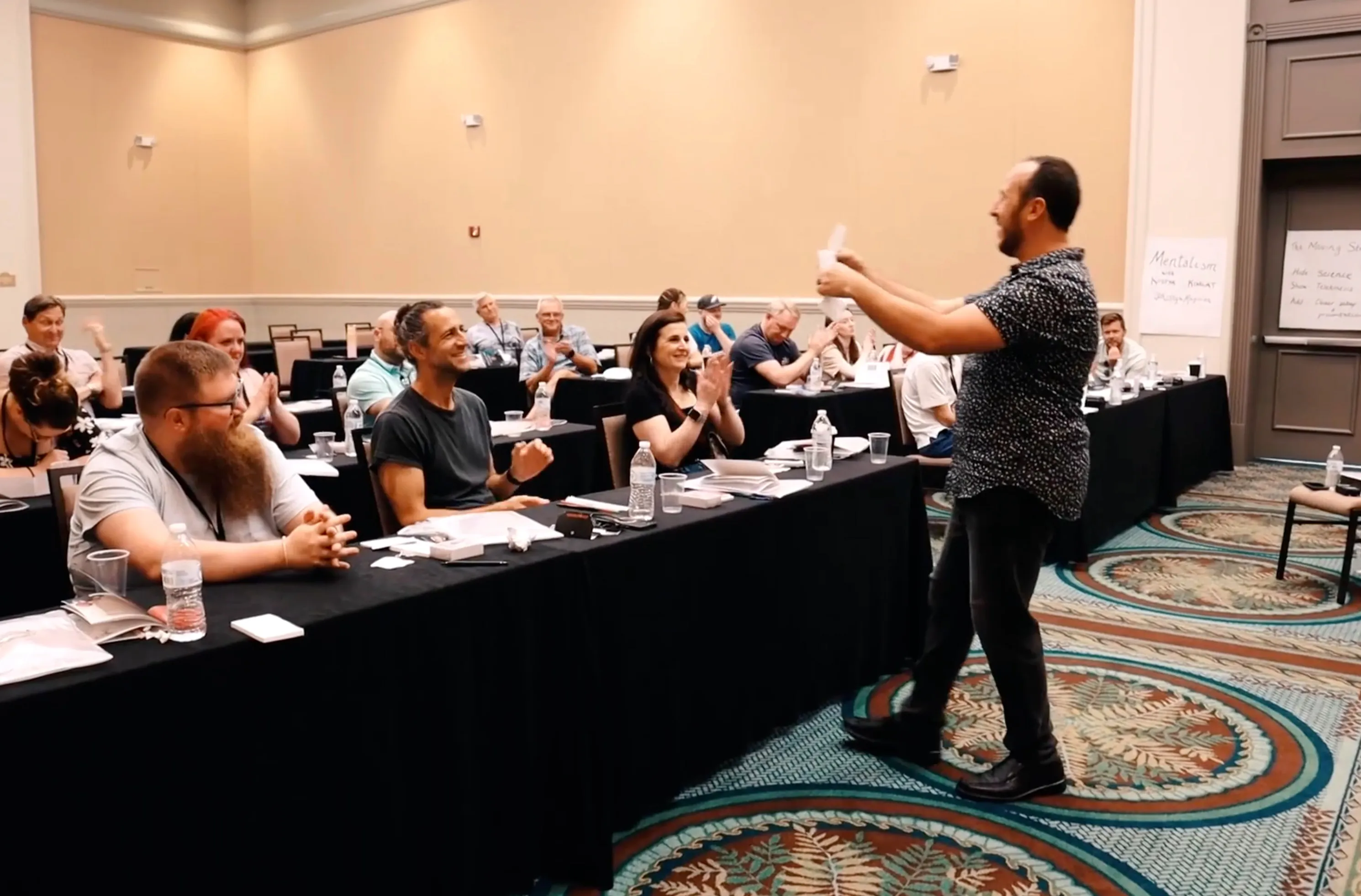 Around fifteen workshop participants at classroom tables applauding as Kostya Kimlat holds up a folded slip of paper at the front of the room