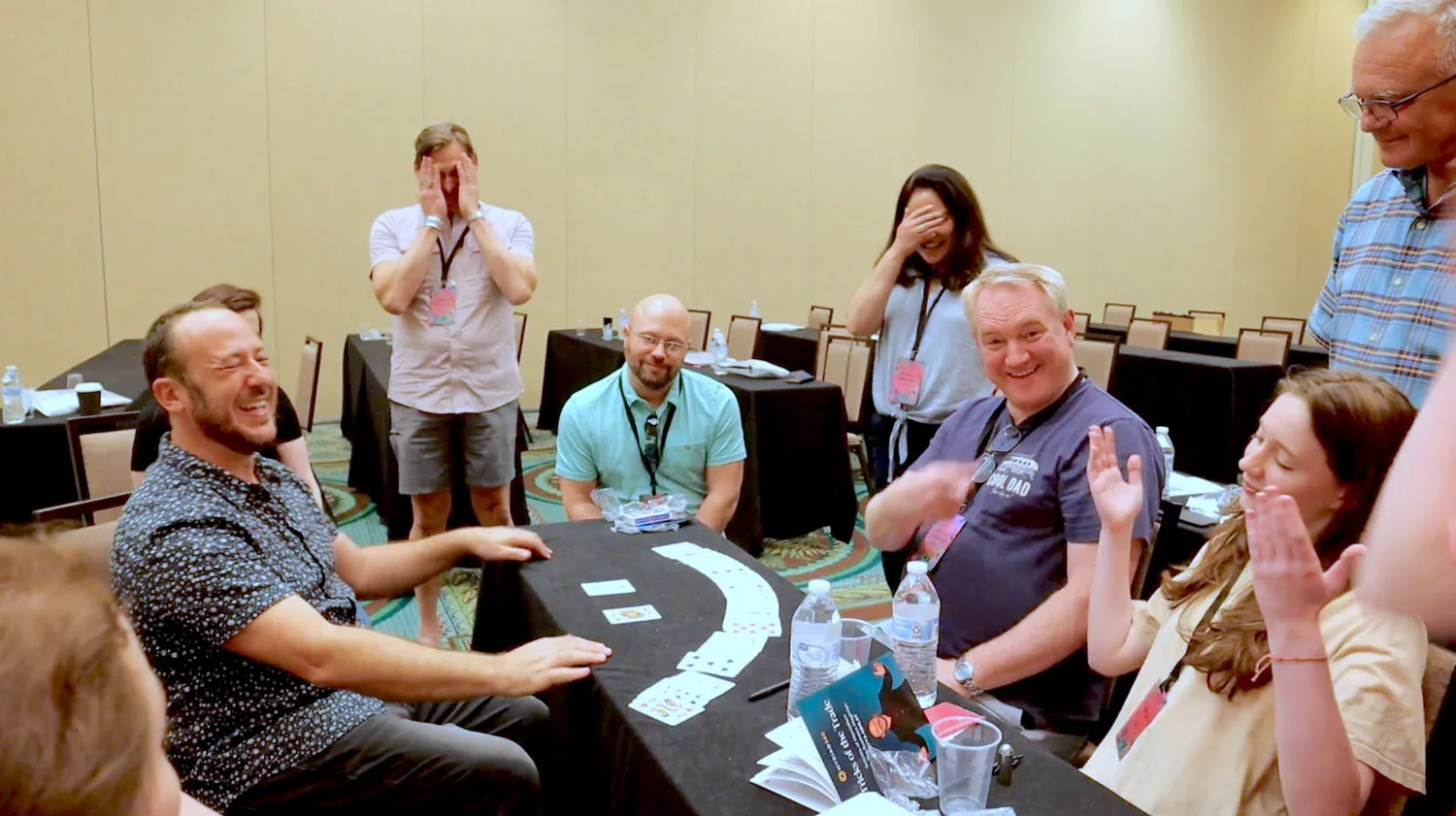 Kostya Kimlat laughing with a conference group around a table as they cover their eyes and applaud, a row of playing cards spread in front of him