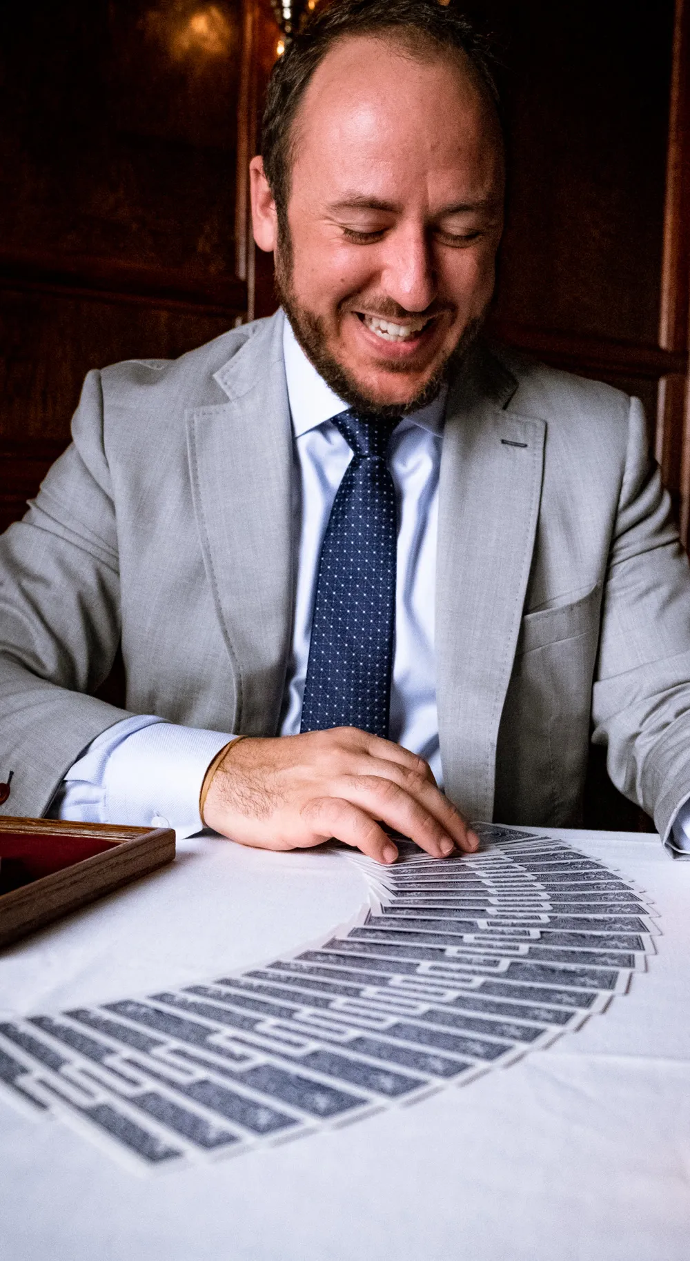 Kostya Kimlat seated at a table, smiling down at a ribbon of playing cards he has spread across white linen