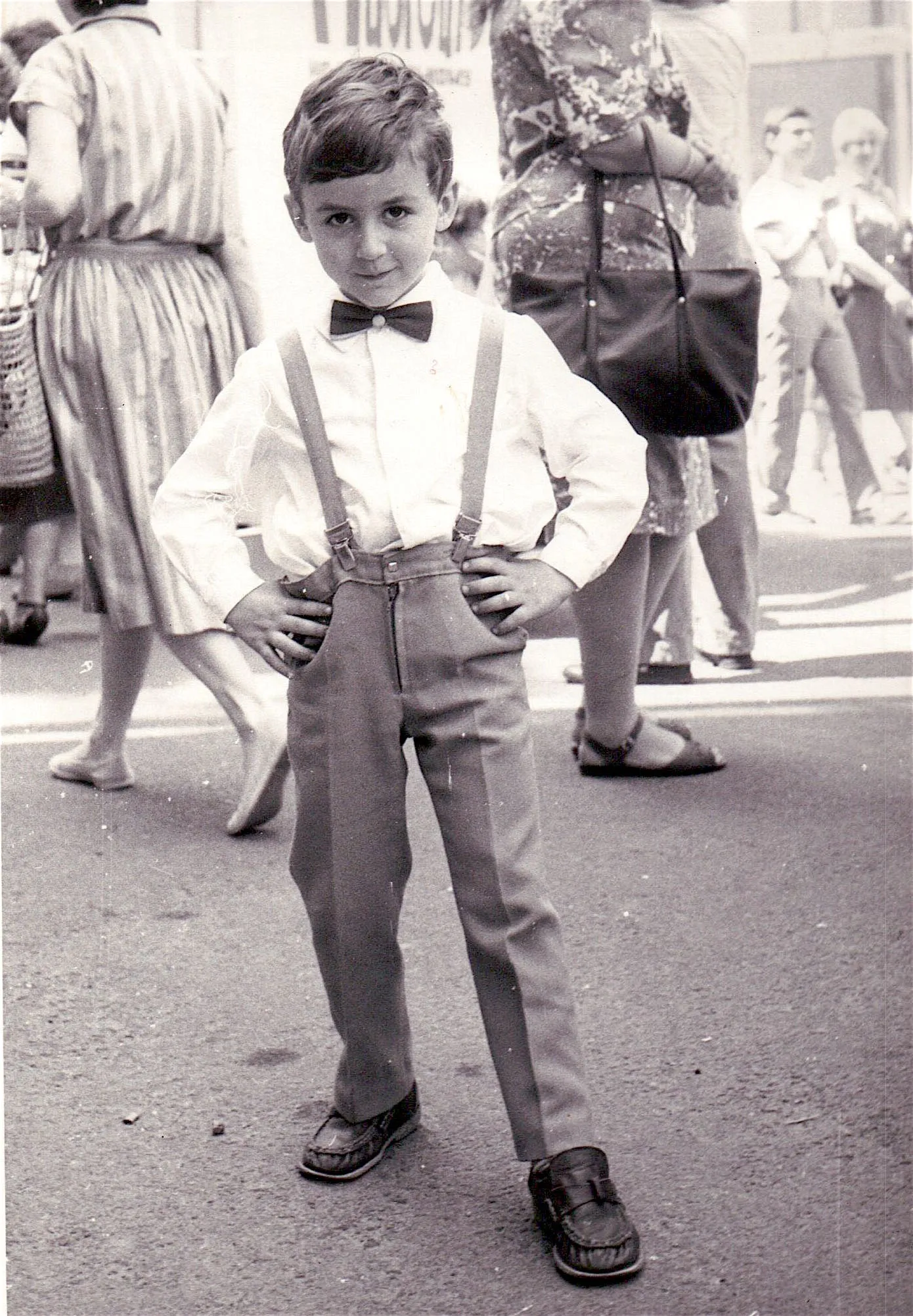 Kostya as a young boy in a bow tie and suspenders, hands on his hips, on a busy street