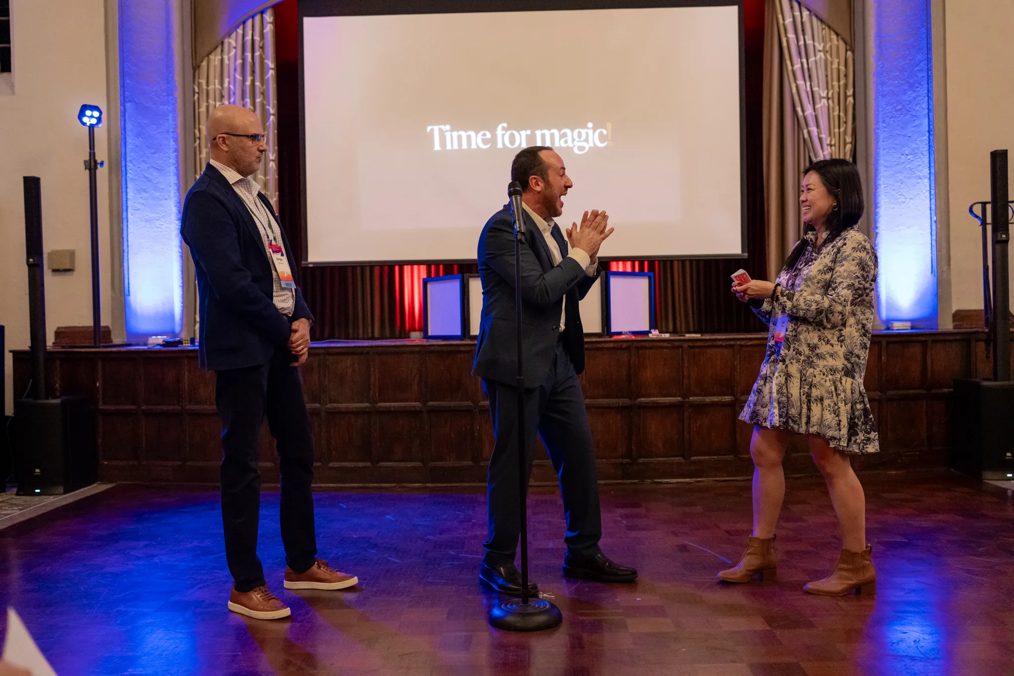 Kostya Kimlat on stage clapping as a volunteer holds a deck of cards, a screen behind them reading “Time for magic”