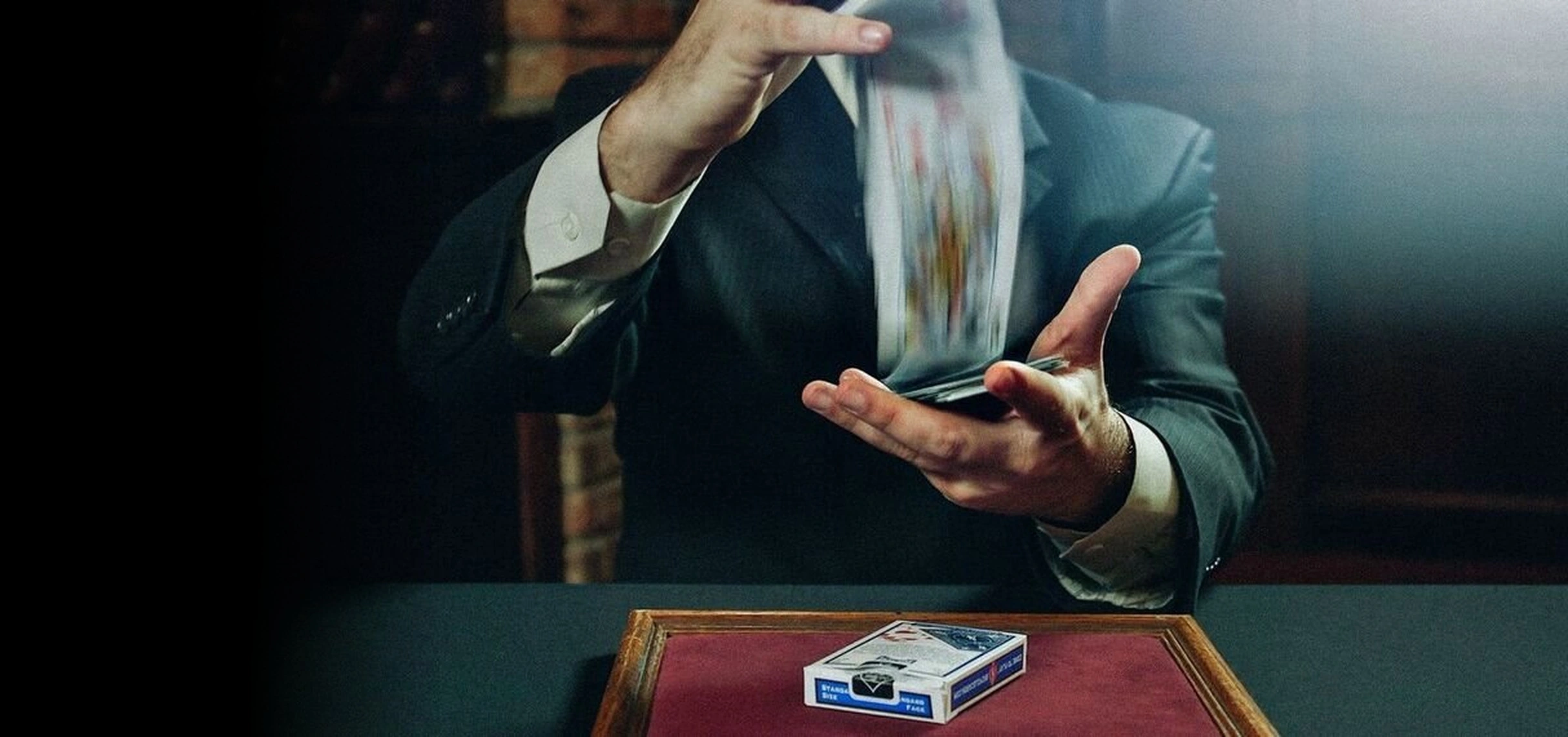 Kostya Kimlat in a suit dribbling a deck of playing cards from one hand to the other above a card case on a green baize table