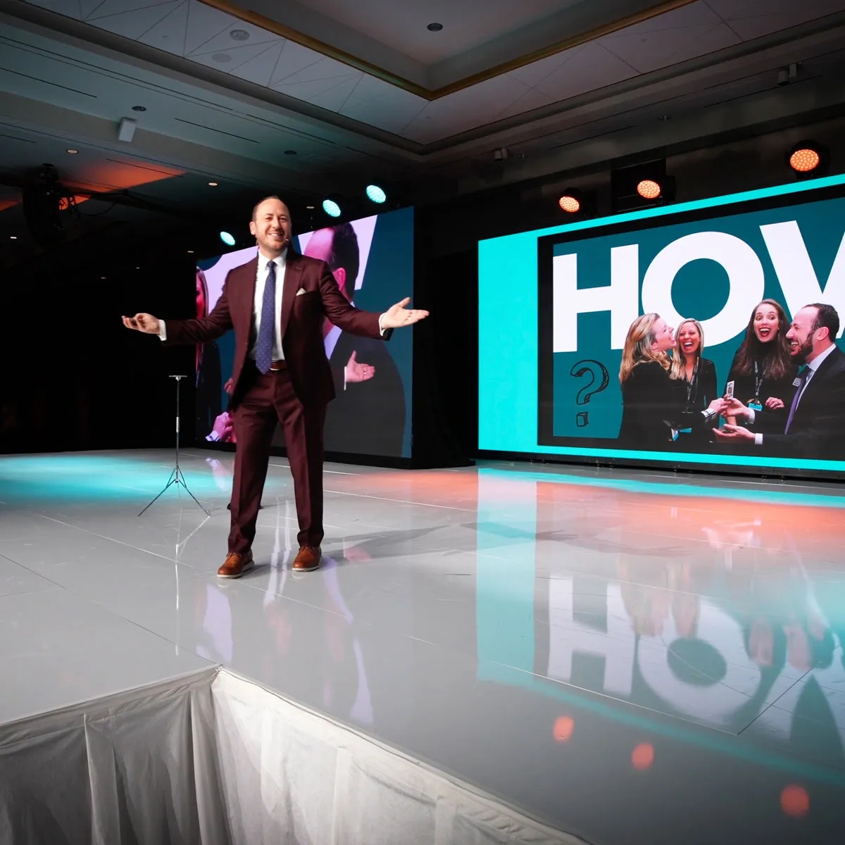 Kostya on a conference stage, arms open, in front of a giant screen showing audience members laughing at a card trick
