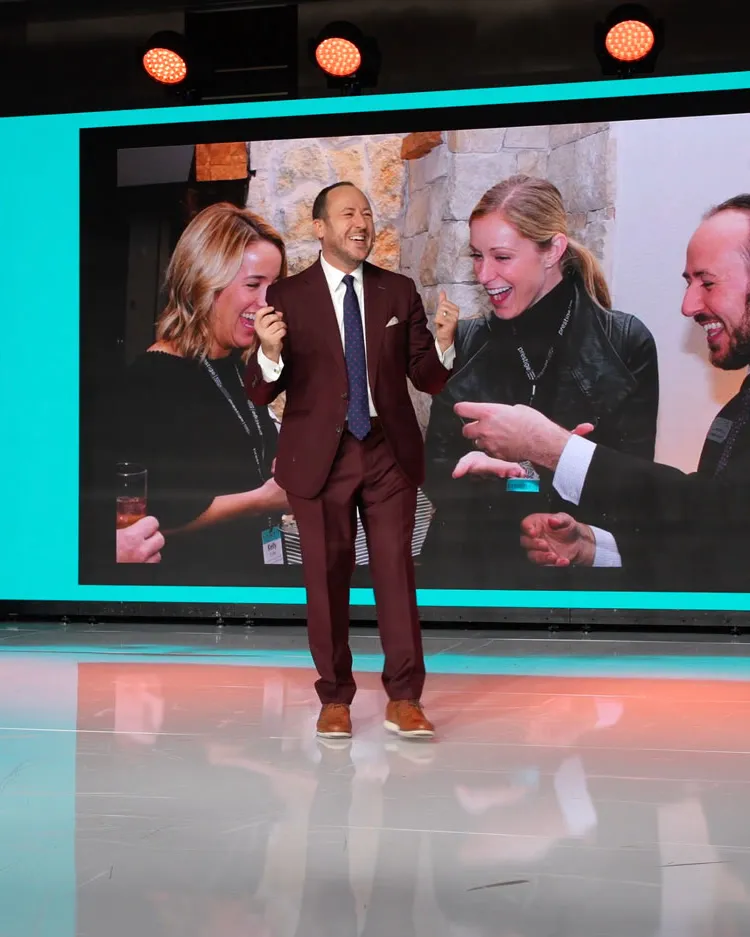 Kostya Kimlat walking downstage in a maroon suit as a large screen behind him shows three guests laughing at a close-up trick