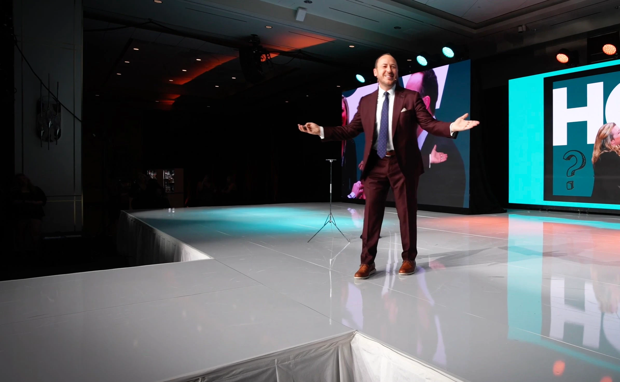 Kostya Kimlat on a ballroom stage with his arms open, beside a large screen reading HOW above a photograph of four people watching a card trick