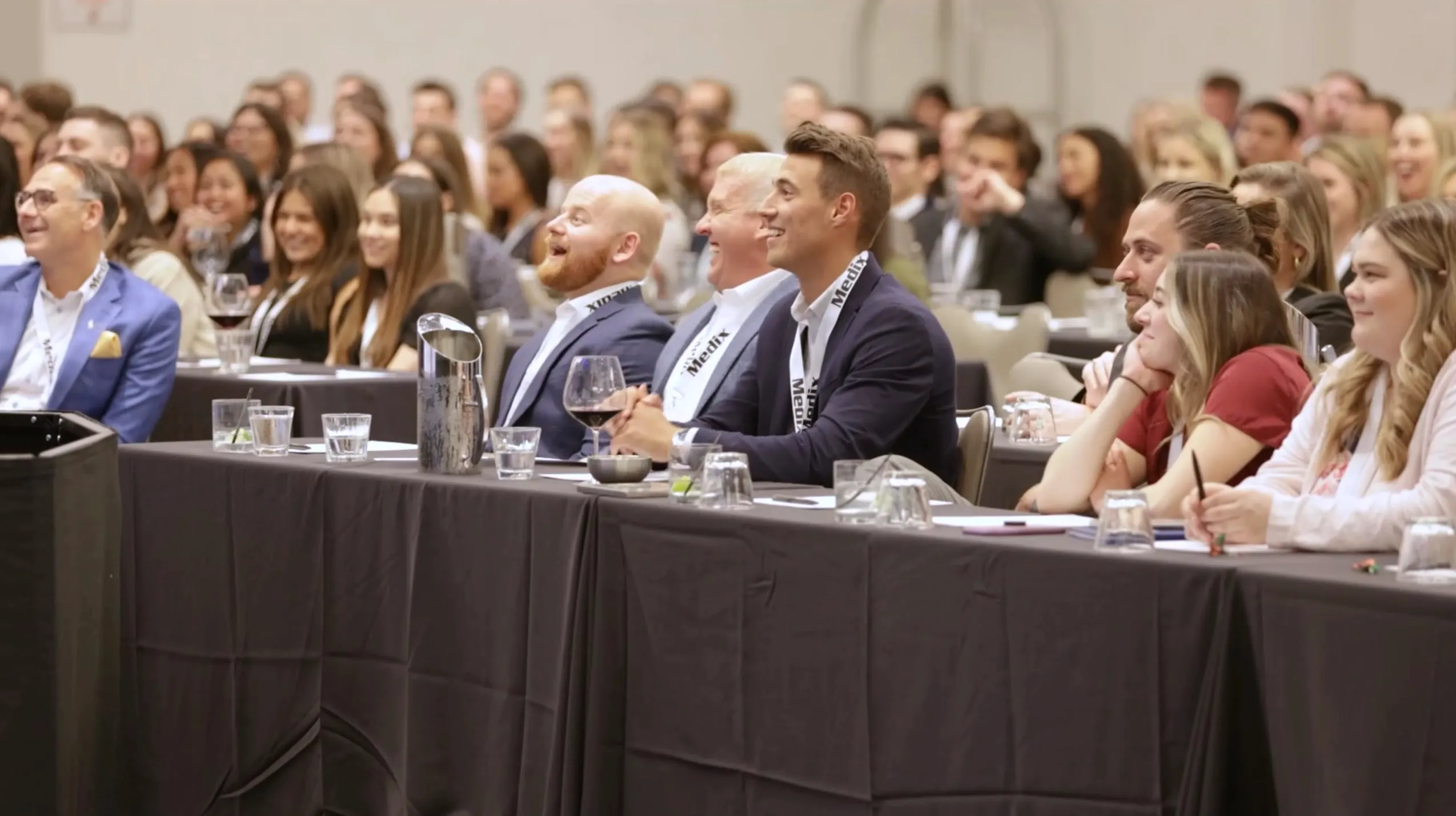 A conference audience at banquet tables laughing together, three men in the front row mid-laugh