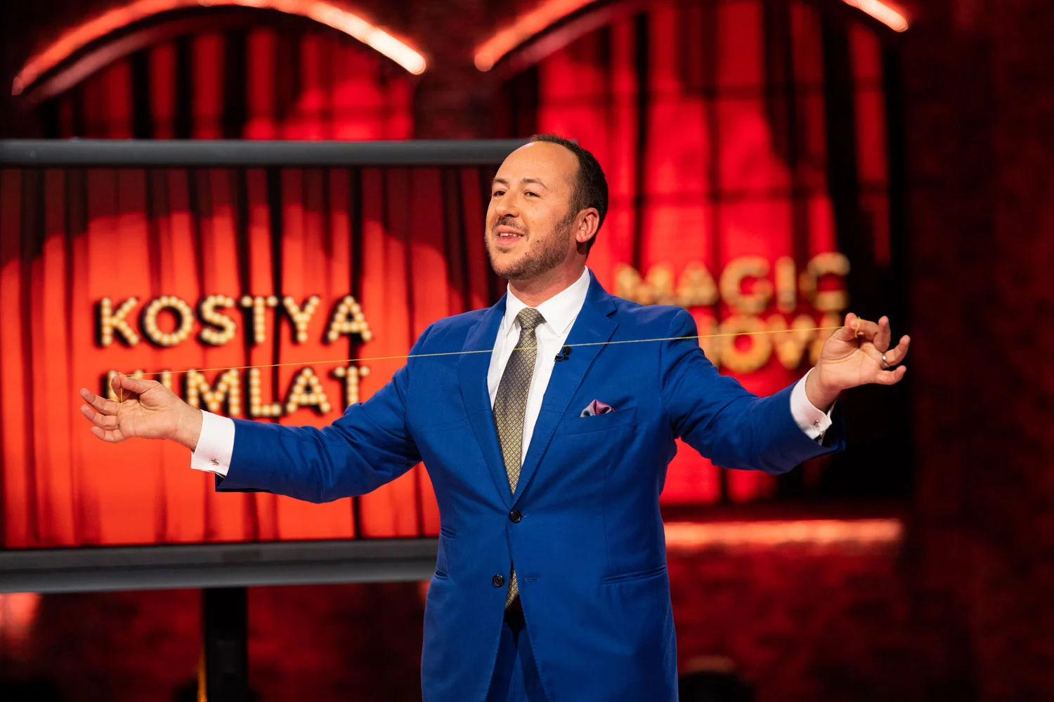 Kostya Kimlat on stage in a blue suit, holding a length of thread taut between his hands