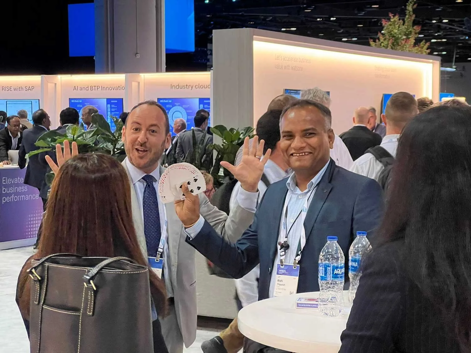 Kostya Kimlat holding a fan of cards beside a laughing attendee with his hand raised, on the SAP Sapphire exhibit floor