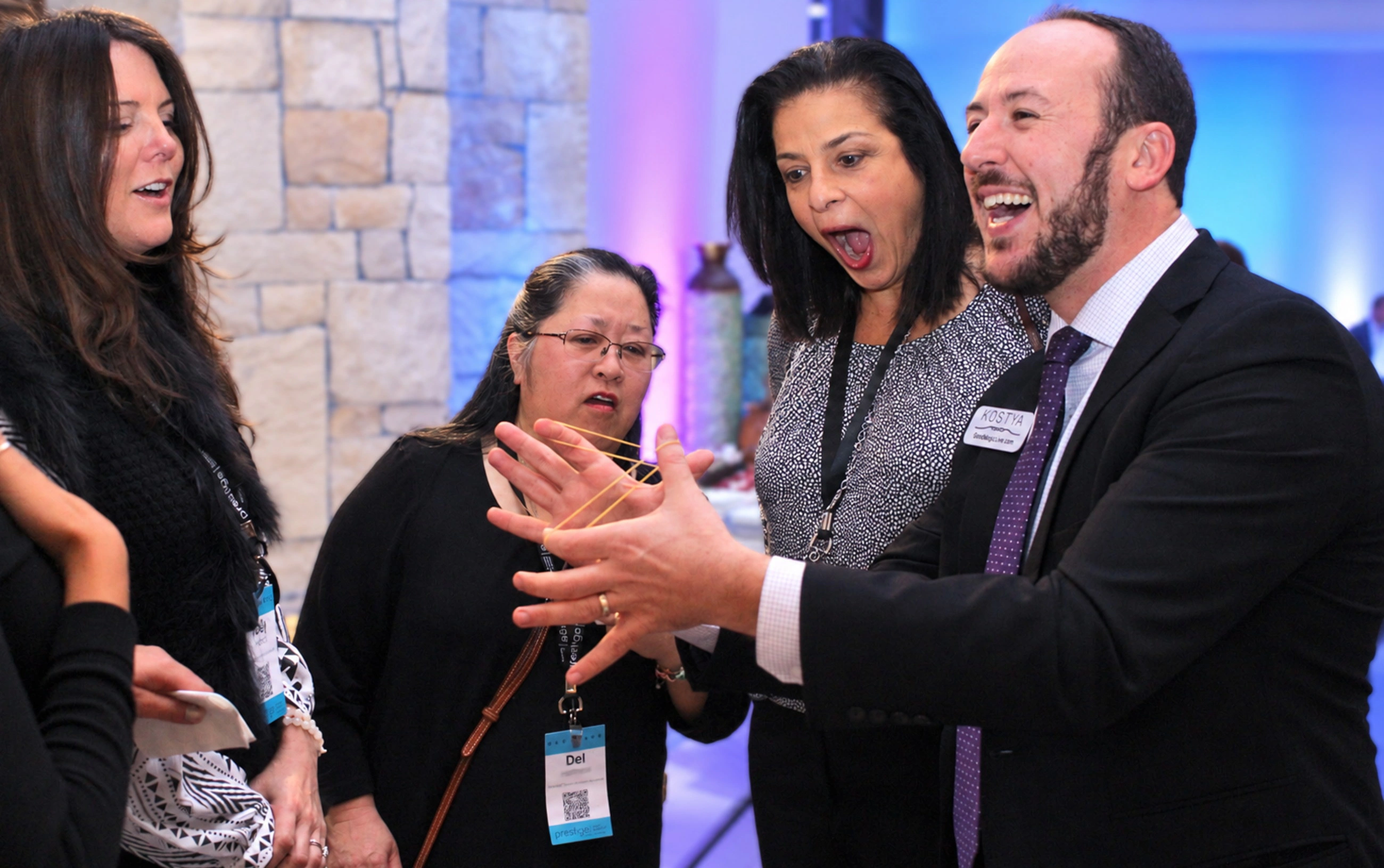 Kostya Kimlat stretching a rubber band between his hands while three guests in name badges react, one open-mouthed in astonishment