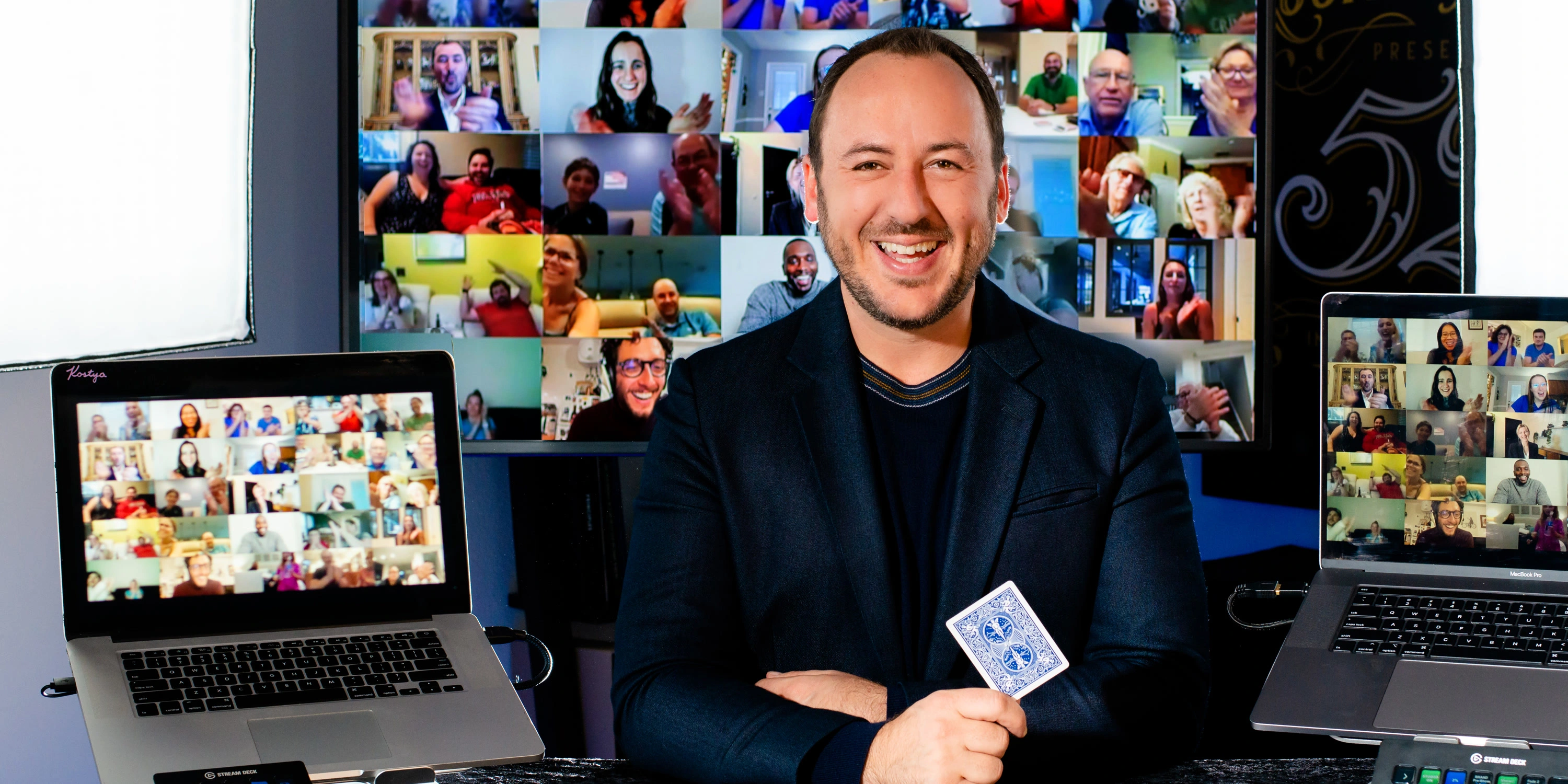 Kostya Kimlat holding a playing card in his studio, a wall of laughing guests on the screen behind him