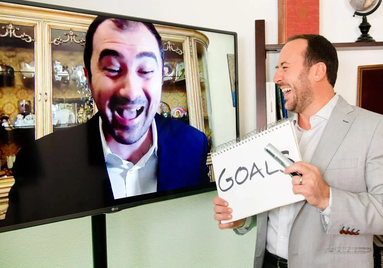 A guest laughing on screen beside Kostya Kimlat holding a hand-drawn sign